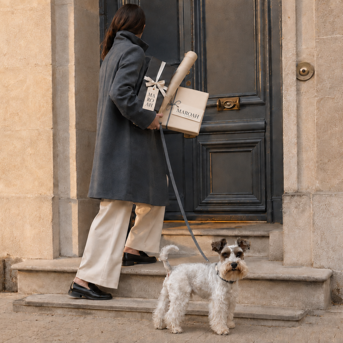 A woman in a dark gray coat and cream trousers stands on the steps of a beige stone building, holding MAROAH gifts. A small white and black dog stands on the steps in front of her.