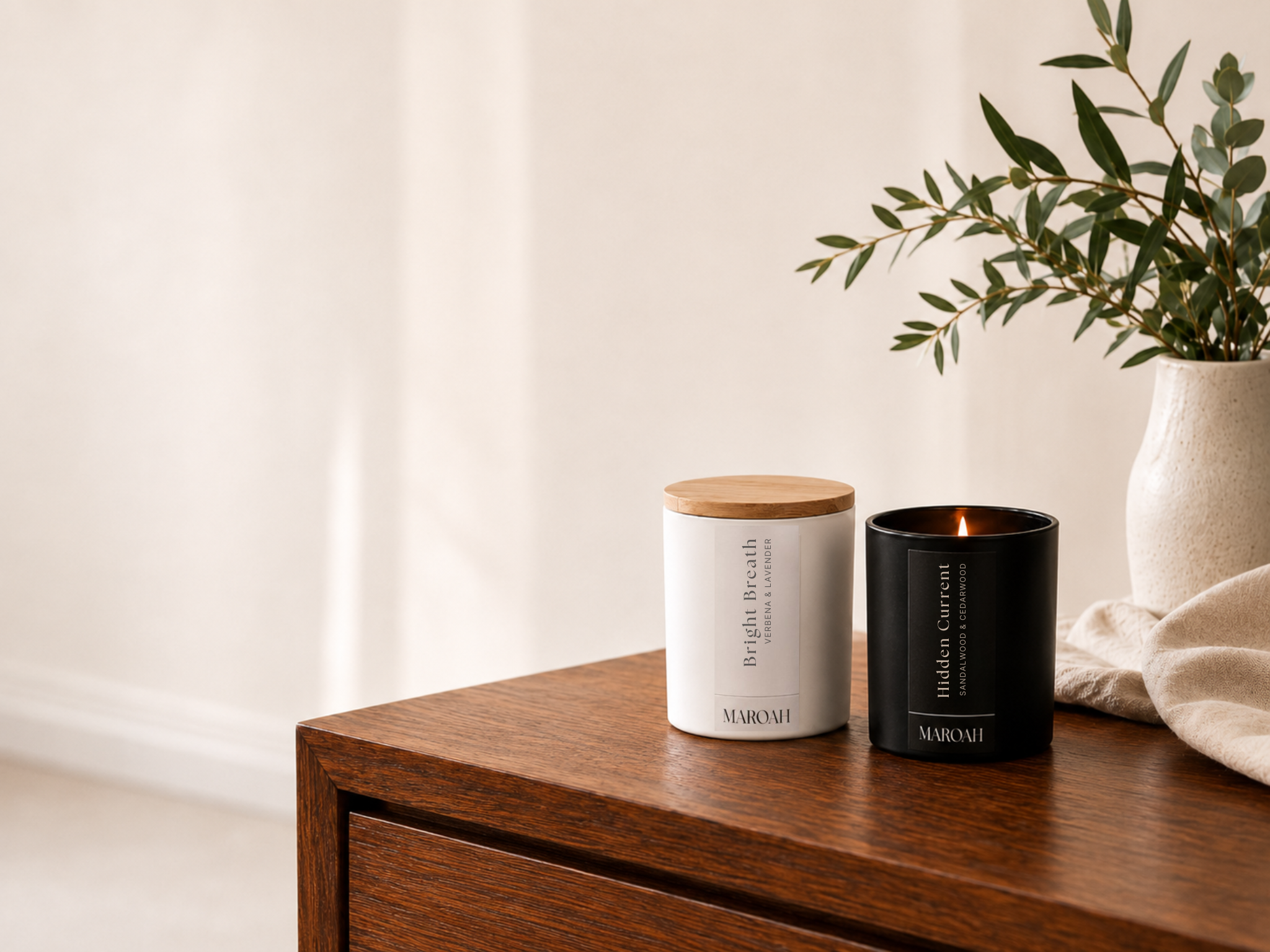 Two MAROAH candles, one white with a light wood lid and one black with vertical text, displayed on a wooden side table next to a vase of greenery.