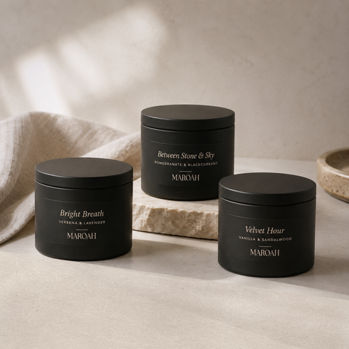 Three matte black MAROAH candle tins arranged on a light stone surface with a draped linen cloth. The tins are labeled Bright Breath, Between Stone & Sky, and Velvet Hour with scent names and notes printed in white and gold text.