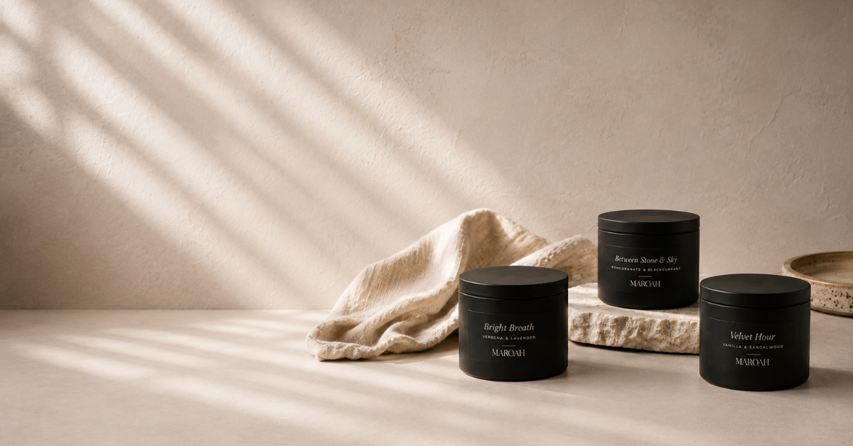 Three MAROAH scented candles in black tins with white text labels, styled on a beige surface with a textured linen cloth and a stone slab beneath, under warm, directional sunlight casting soft shadows.